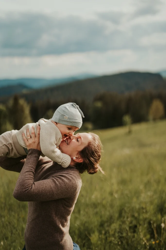 Recenze – rodinné focení: maminka drží miminko v náručí na louce, přirozený moment a emoce s horami v pozadí.