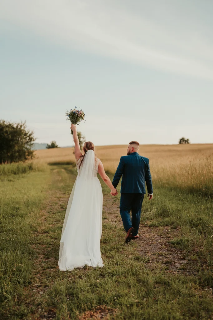 Recenze „Svatební focení – Lenka“ – novomanželé jdou po cestě mezi poli, nevěsta drží kytici v jemném večerním světle.