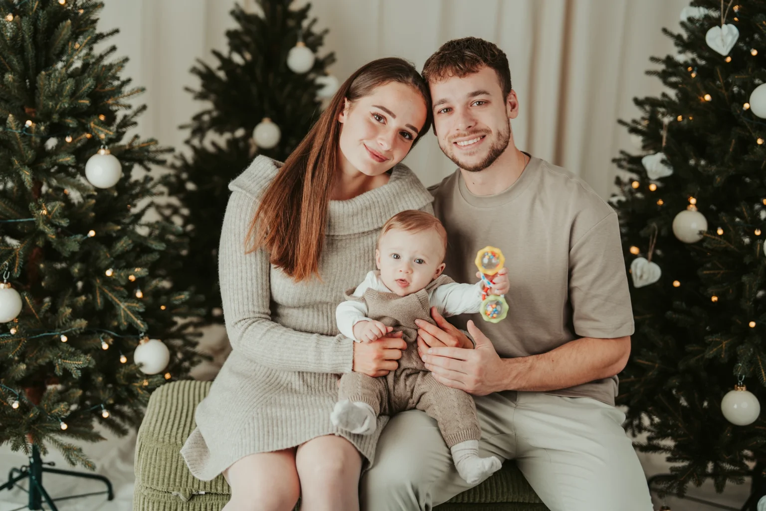 Maminka a tatínek sedí s miminkem mezi dvěma vánočními stromky.
